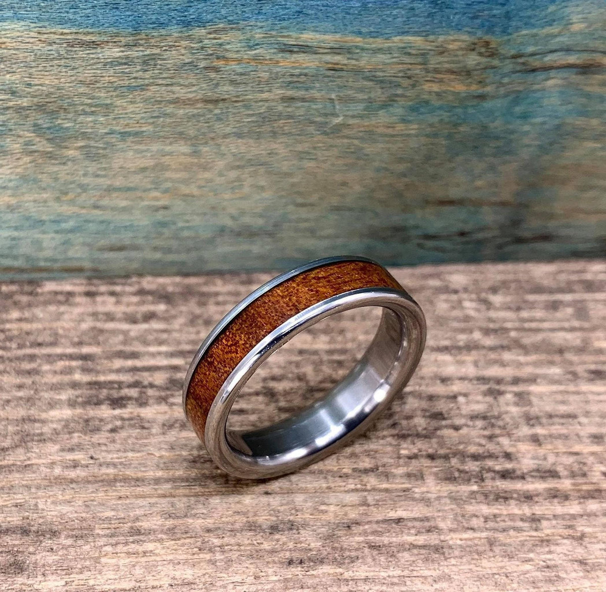 A silver-colored ring with a brown wood inlay is angled on a wooden surface, with a blue-green background.