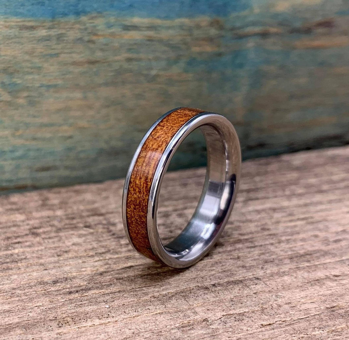 A silver-colored ring with a brown wood inlay is angled on a wooden surface, with a blue-green background.