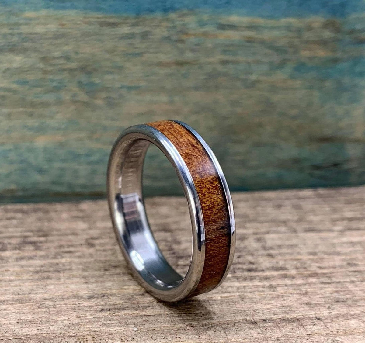 A silver-colored ring with a brown wood inlay is angled on a wooden surface, with a blue-green background.