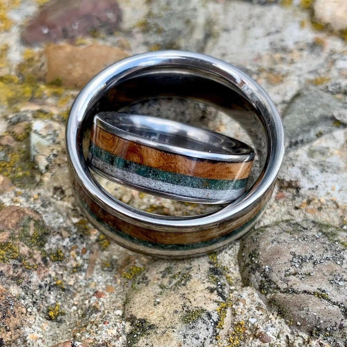 A wide silver ring with wood, green, and cream inlay, with a narrower matching ring nested inside, on a rocky surface.