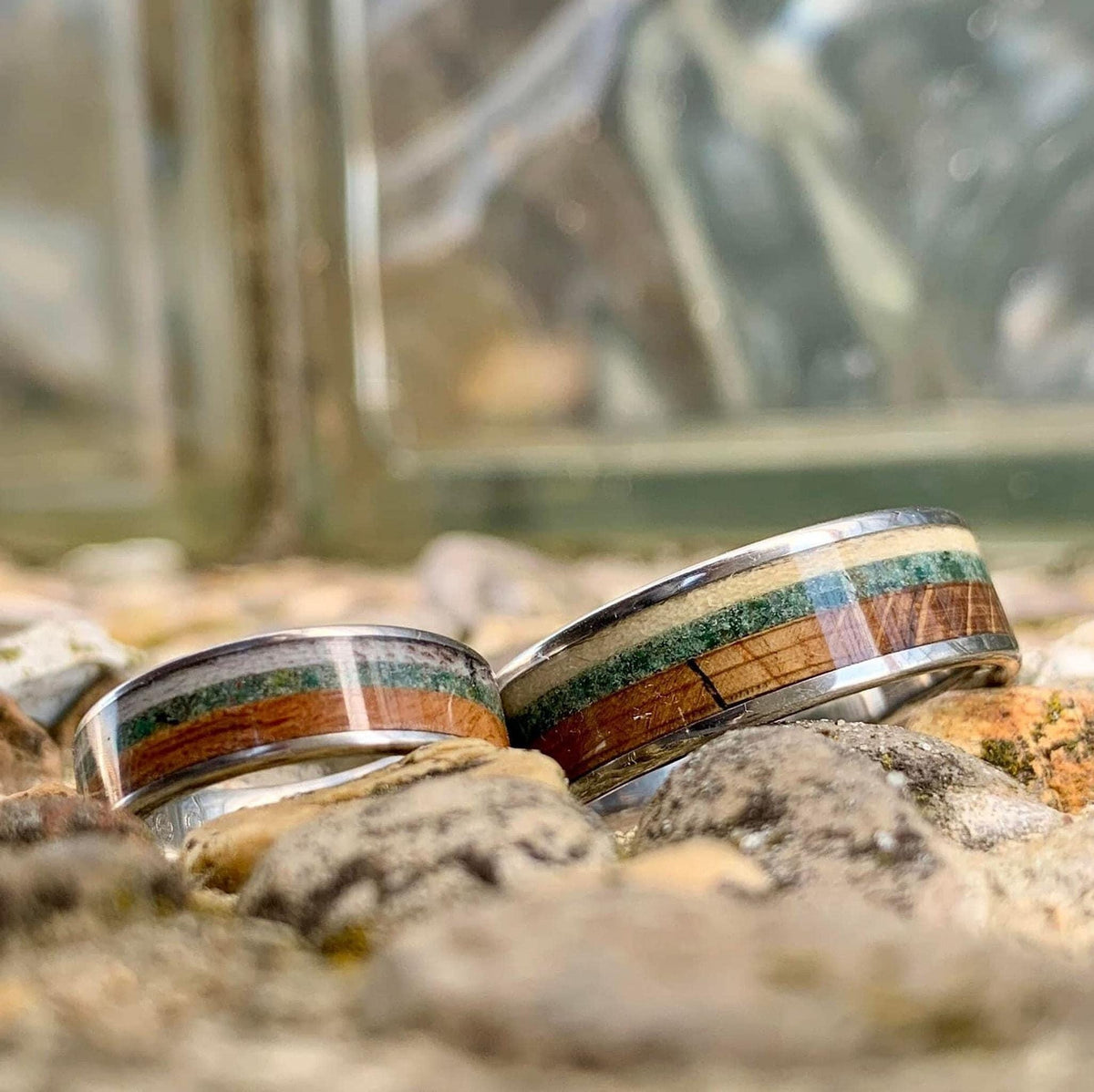 Two silver rings with wood, green, and cream inlay, one wider than the other, resting on a gravelly surface.