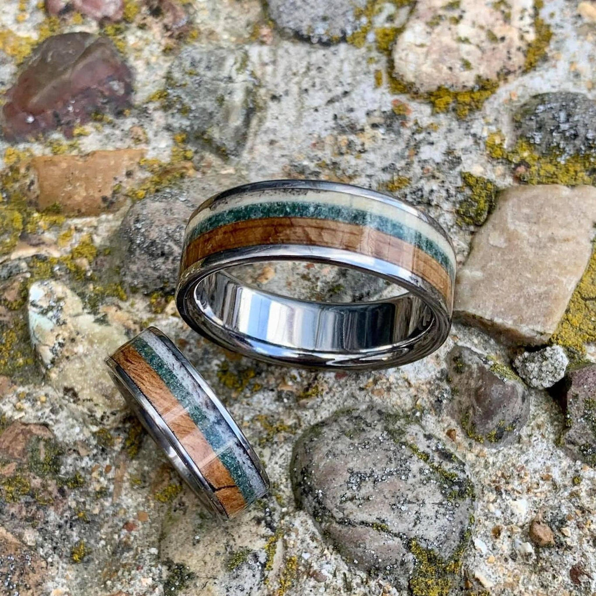 A wide silver ring with wood, green, and cream inlay, with a narrower matching ring beside it, on a rocky surface.