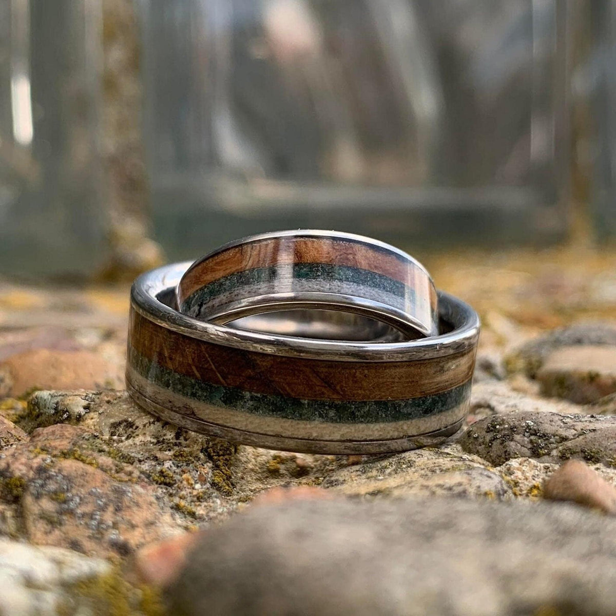 Two silver rings with wood, green, and cream inlay, one wider than the other, with the narrower ring leaning against the wider one.