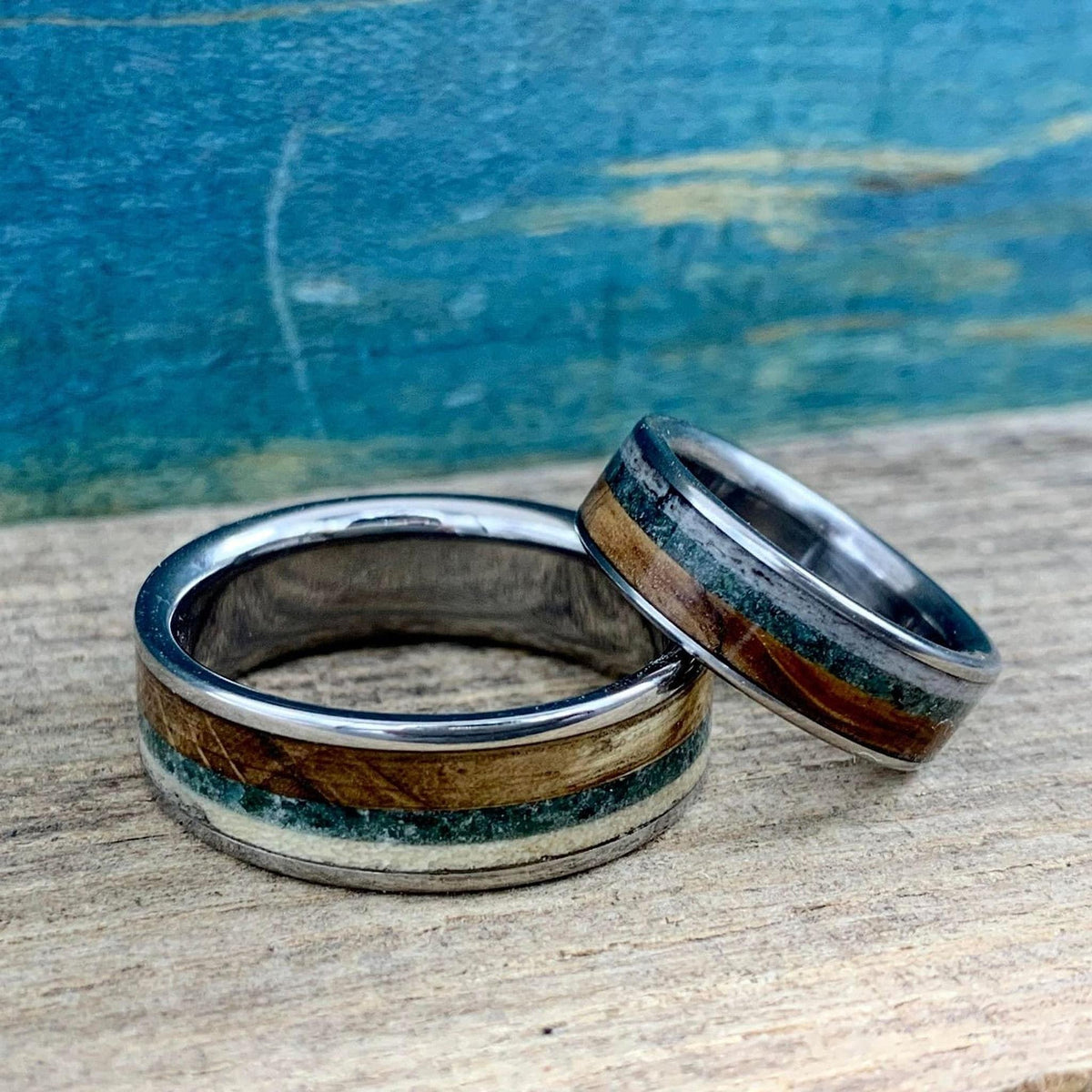 Two silver rings with wood, green, and cream inlay, one wider than the other, resting on a wooden surface.