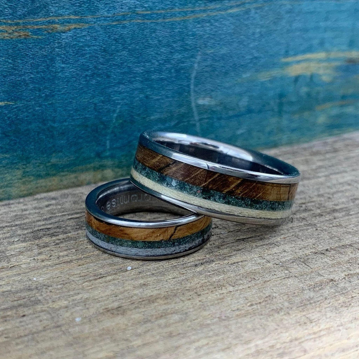 Two silver rings with wood, green, and cream inlay, one wider than the other, resting on a wooden surface with a blue background.