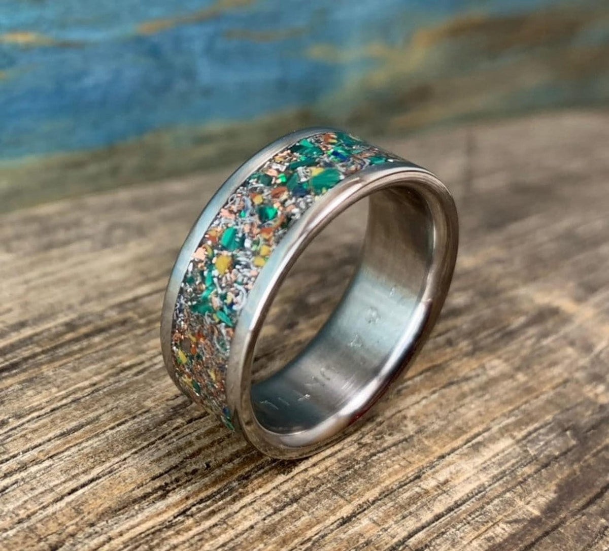 A silver-colored ring with a wide, colorful inlay of green, orange, and brown fragments, on a wooden surface.