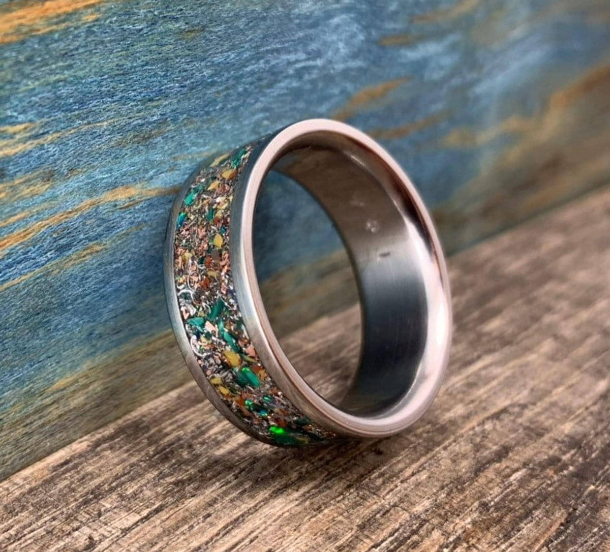 A silver-colored ring with a colorful inlay of green, orange, and brown fragments, angled on a wooden surface.