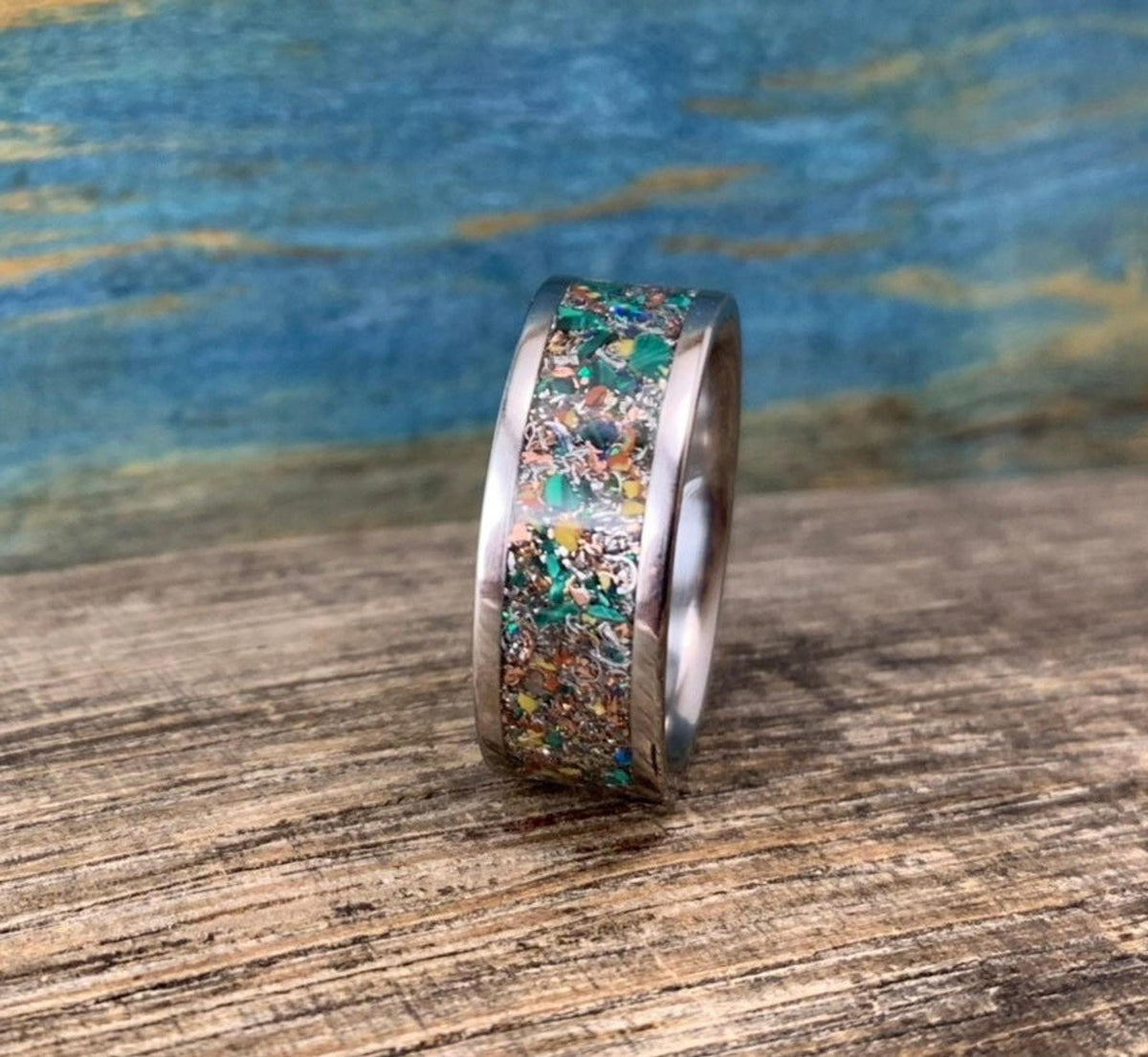 A silver-colored ring with a colorful inlay of green, orange, and brown fragments, viewed from a slight angle on a wooden surface.