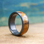 A black titanium ring with a light brown wood inlay, featuring a visible wood grain, is angled on a wooden surface.