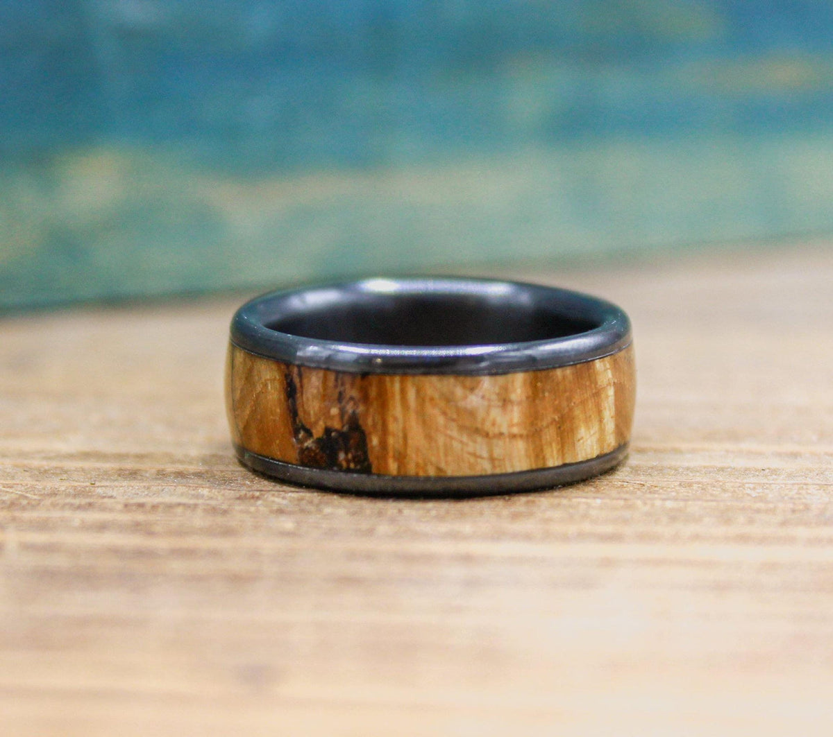 A black titanium ring with a light brown wood inlay is viewed from a slightly elevated angle, showing the top and inner band.