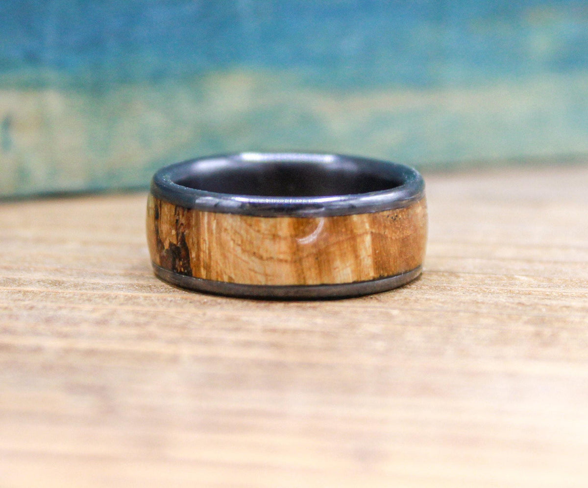 A black titanium ring with a light brown wood inlay is centered on a wooden surface, showing its full width.