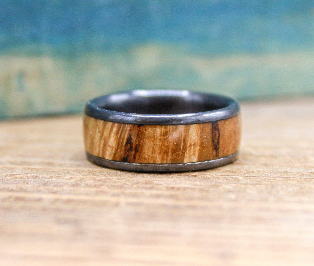 A black titanium ring with a light brown wood inlay, showcasing its texture and color variations, is on a wooden surface.