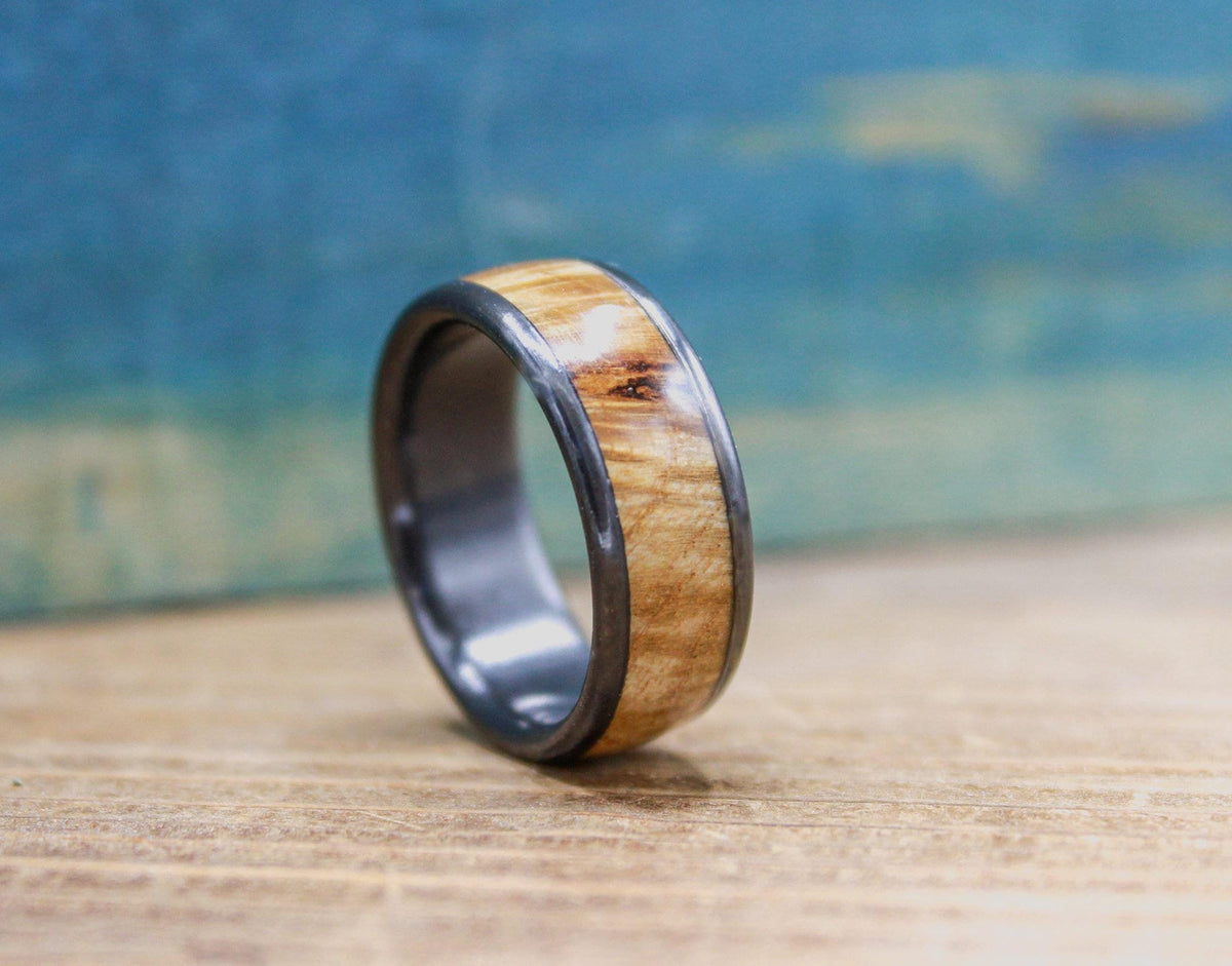 A black titanium ring with a light brown wood inlay, exhibiting a distinct wood grain, is angled on a wooden surface.