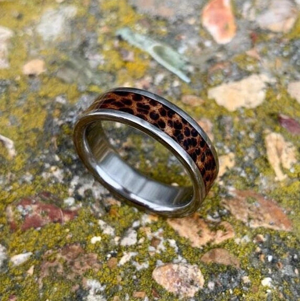 An angled view of the titanium wedding band featuring a patterned red palm wood inlay on a textured stone surface.