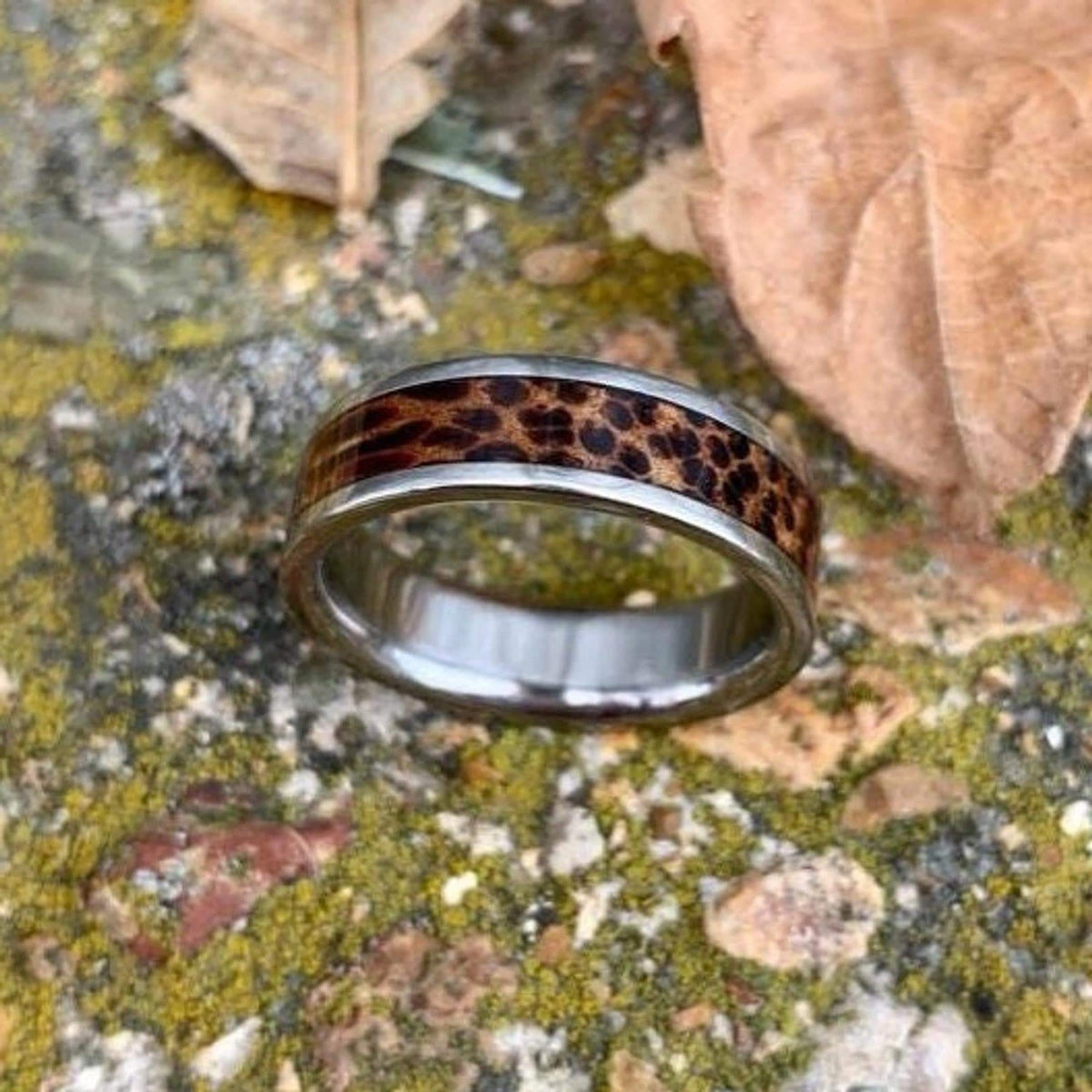 A titanium ring with a dark red palm wood inlay sits on a mottled grey stone and leaf background.