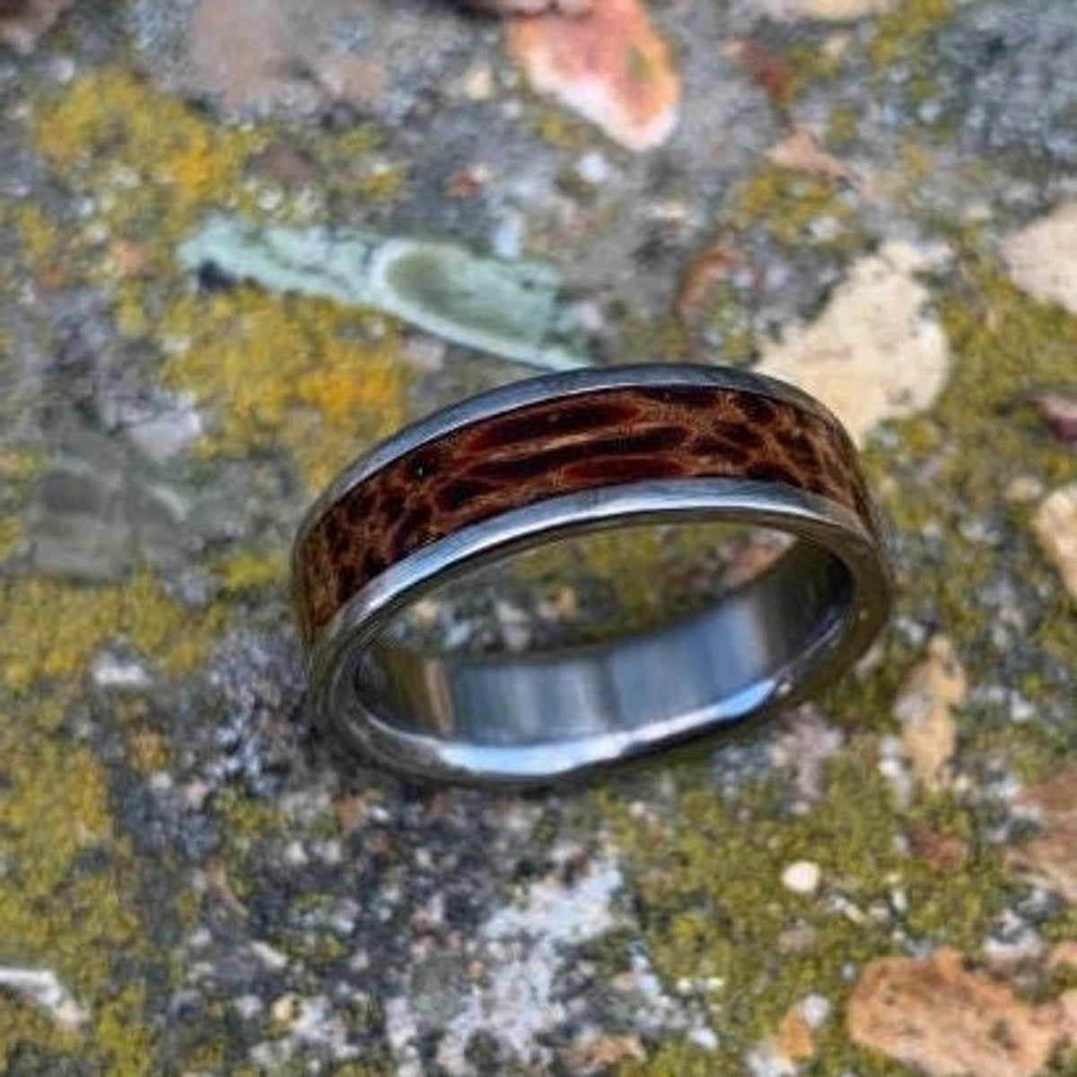 A titanium ring with a distinctive red palm wood inlay displayed on a mossy stone backdrop.