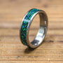 A silver-colored ring with a green and blue speckled inlay is angled on a wooden surface.