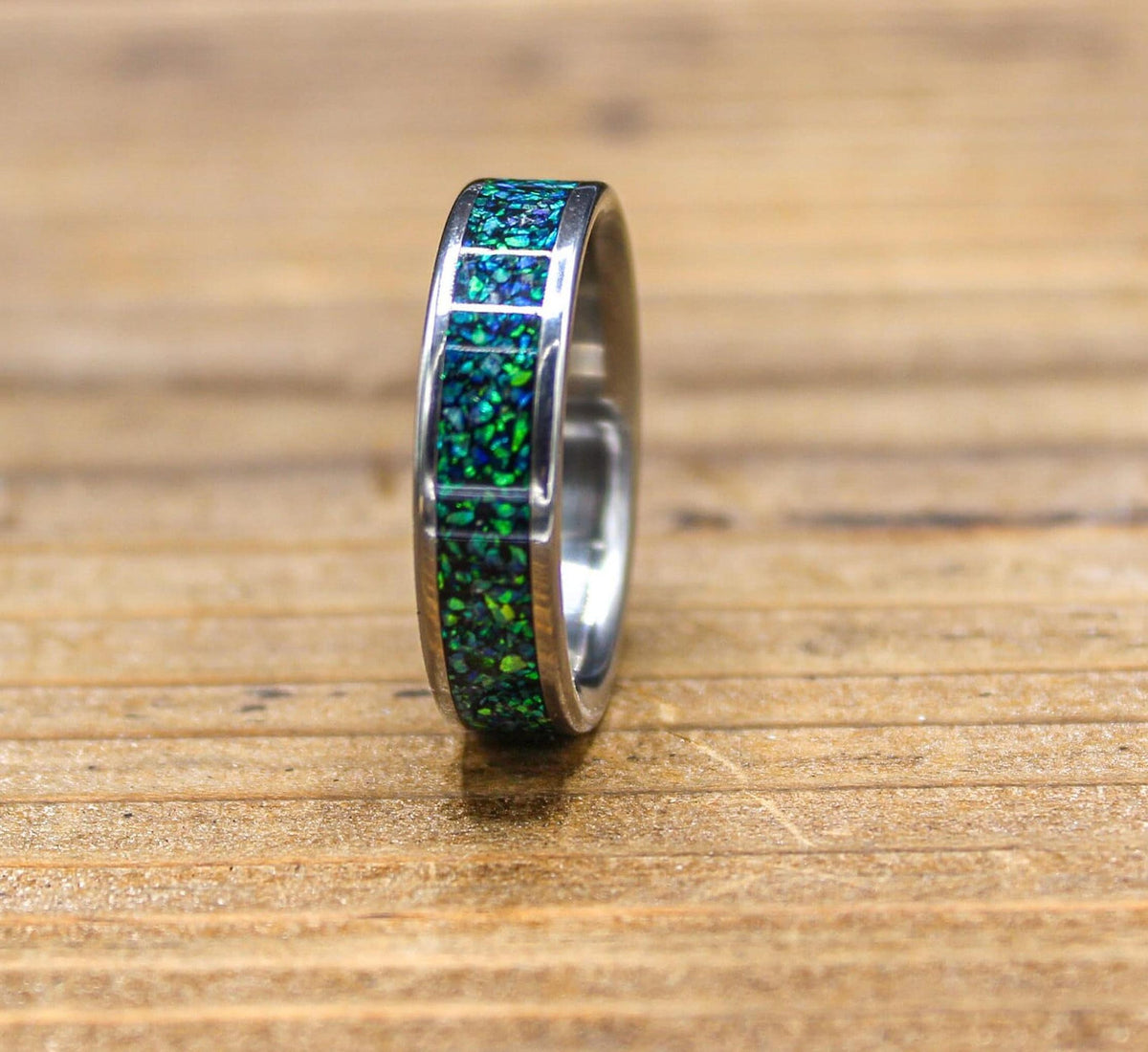 A silver-colored ring with a green and blue speckled inlay is shown from a side view on a wooden surface.