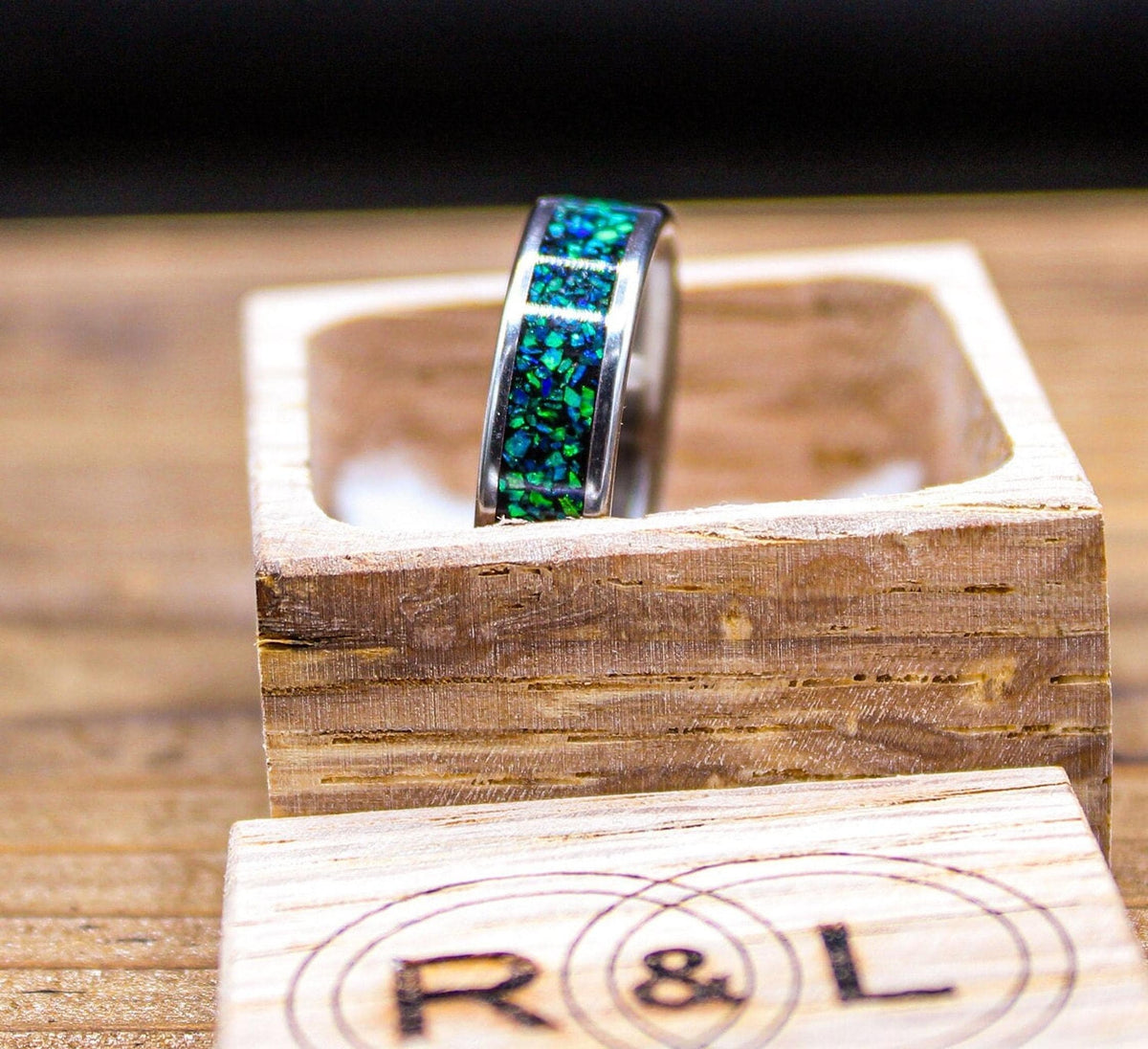 A silver-colored ring with a green and blue speckled inlay stands upright inside an open light wood box with a circular R&L logo.
