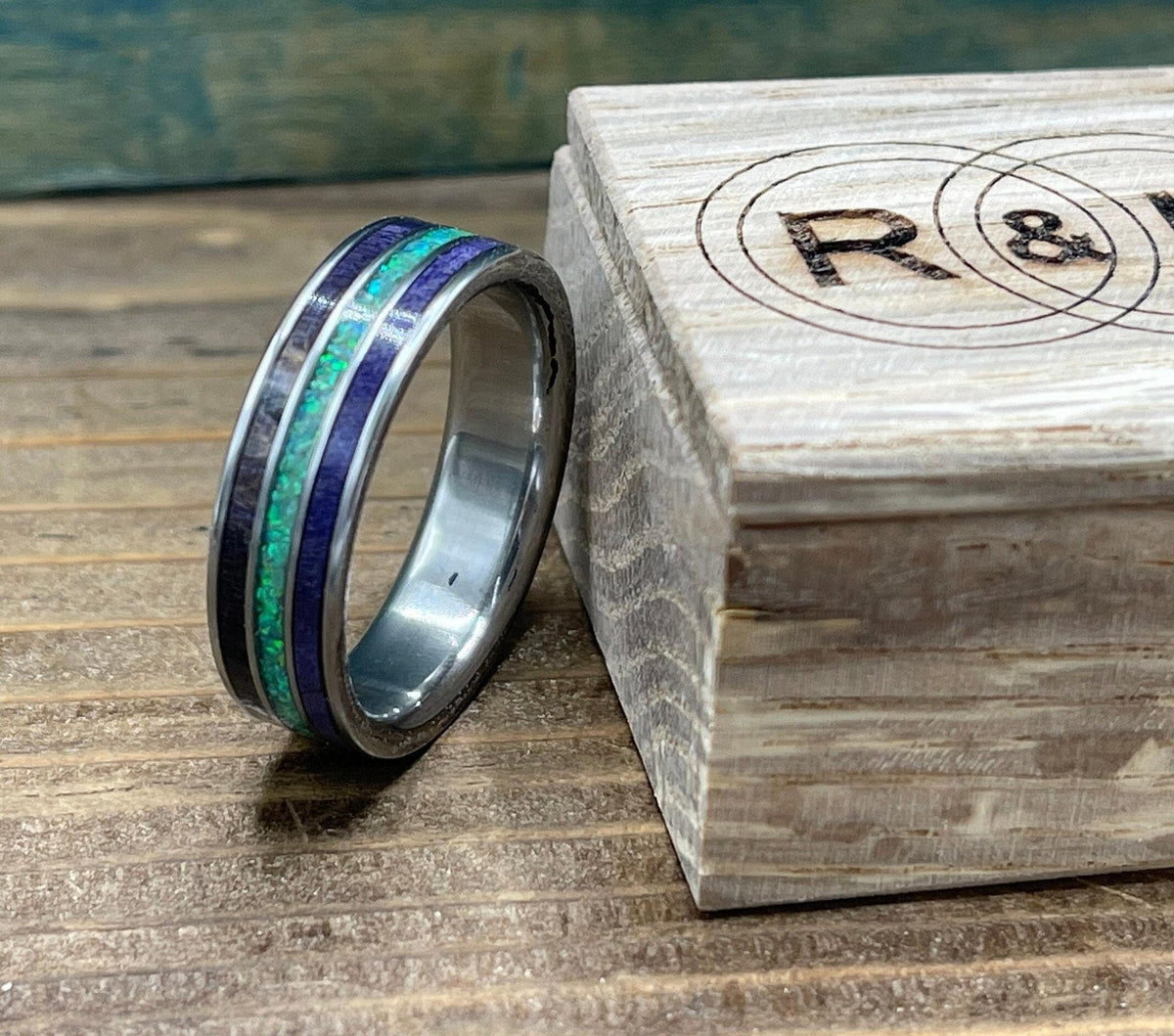 A silver ring with purple wood and green opal inlay rests against a light wooden box with a circular "R & L" logo.