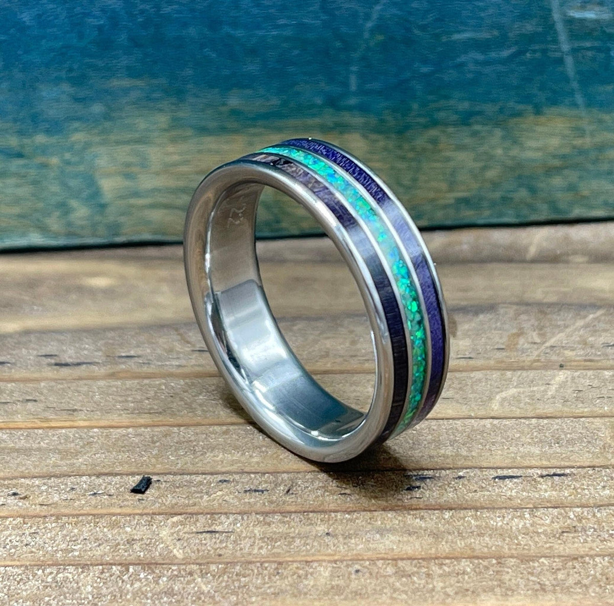 A silver ring with purple wood and green opal inlay is positioned on a wooden surface, with a blue-green background.
