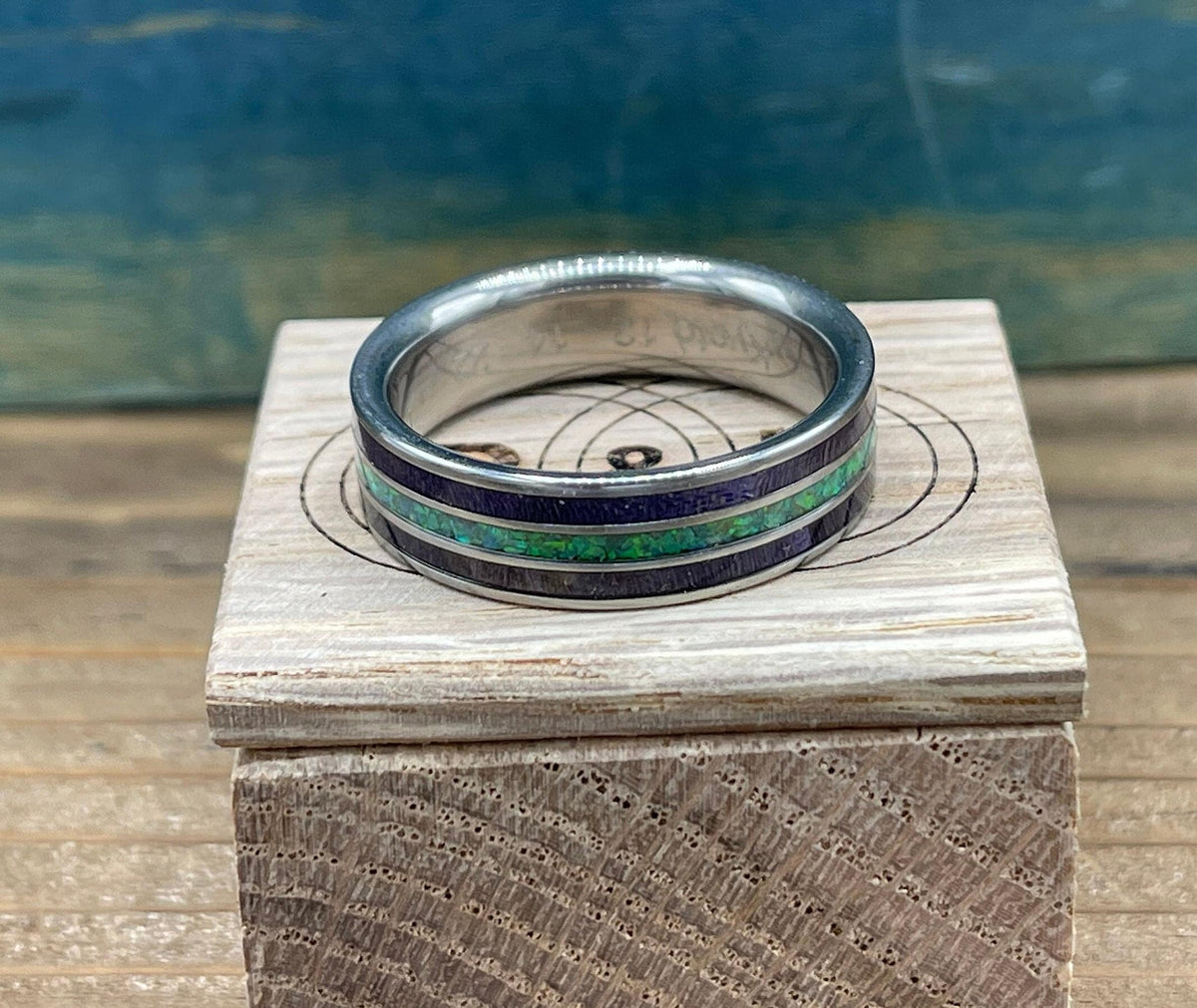 A silver ring with purple wood and green opal inlay sits on a light wooden box with a circular target-like design.