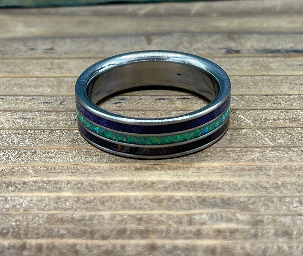 A silver ring with purple wood and green opal inlay is shown from a top-down perspective on a wooden surface.