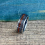 A silver-colored ring with a wide, textured gray band and a thin, reddish-brown stripe running through its center, viewed from a slight angle on a wooden surface.
