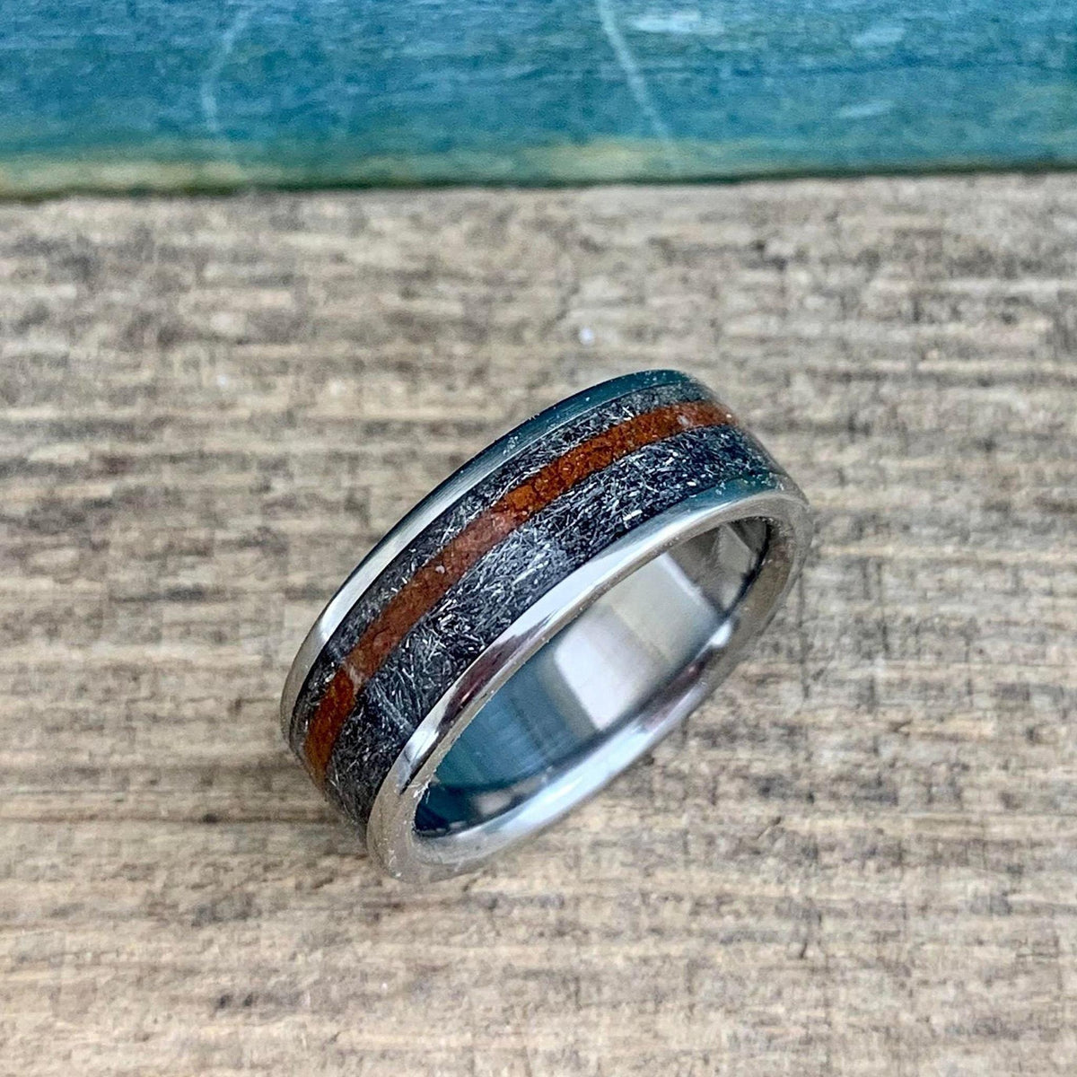 A silver-colored ring with a textured gray band and a reddish-brown stripe, viewed from a slight angle on a wooden surface with a blue background.