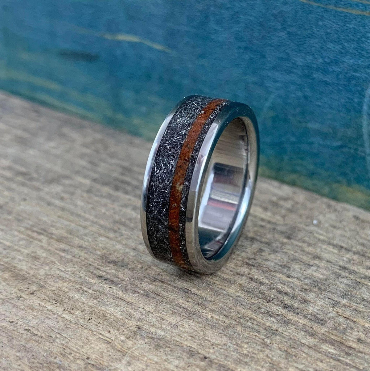 A silver-colored ring with a textured gray band and a reddish-brown stripe, viewed from a slight angle on a wooden surface with a blue background.