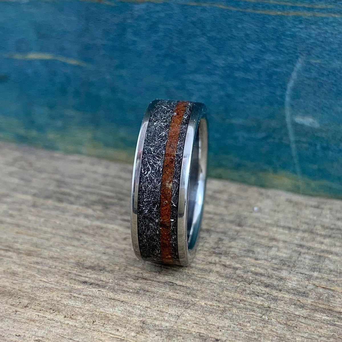 A silver-colored ring with a textured gray band and a reddish-brown stripe, viewed head-on on a wooden surface with a blue background.