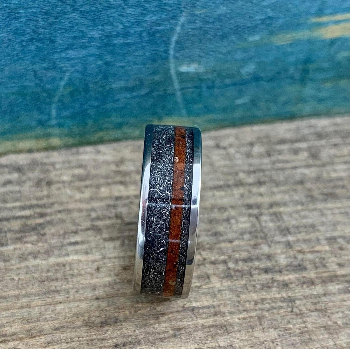 A silver-colored ring with a textured gray band and a reddish-brown stripe, viewed head-on on a wooden surface with a blue background.