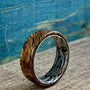 A wooden ring with a silver-colored interior band featuring an engraved floral pattern, resting on a wooden surface with a blue background.