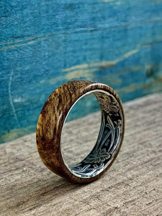 A wooden ring with a silver-colored interior band featuring an engraved floral pattern, resting on a wooden surface with a blue background.