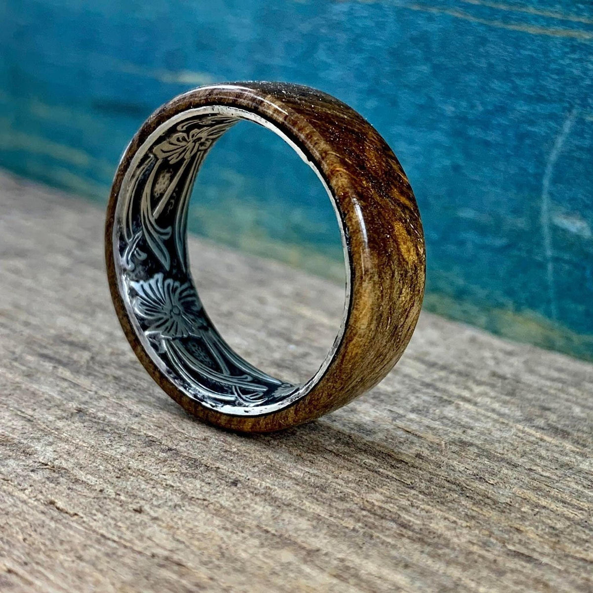 A wooden ring with a silver-colored interior band, engraved with a floral design, is angled on a wooden surface against a blue backdrop.