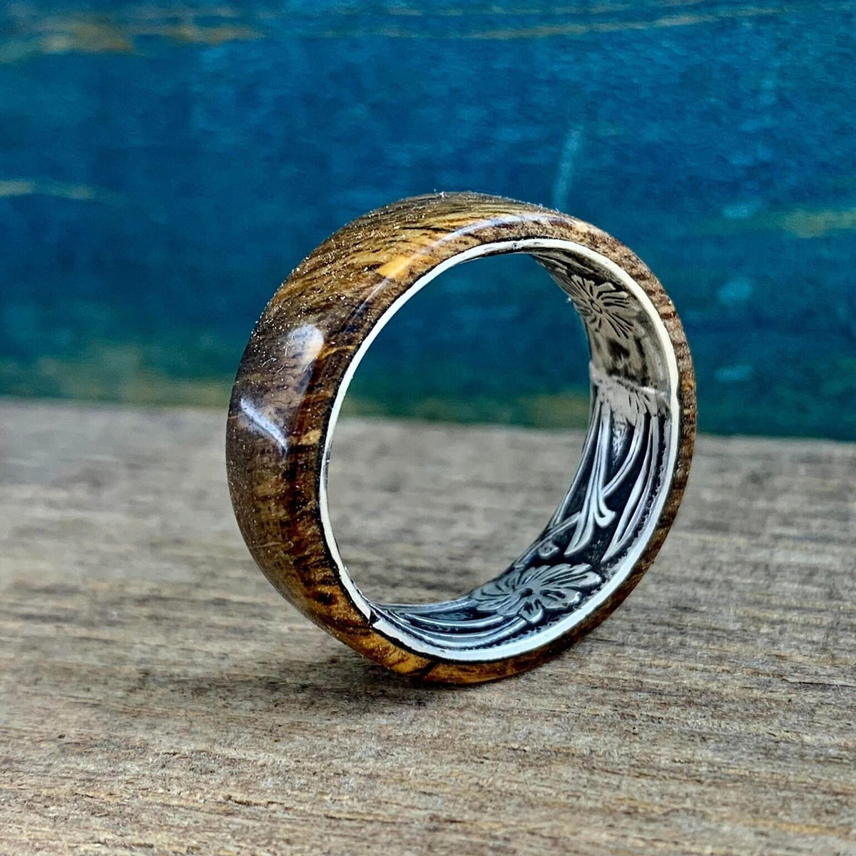 A wooden ring with a silver-colored interior band, engraved with a floral pattern, is shown at an angle on a wooden surface.