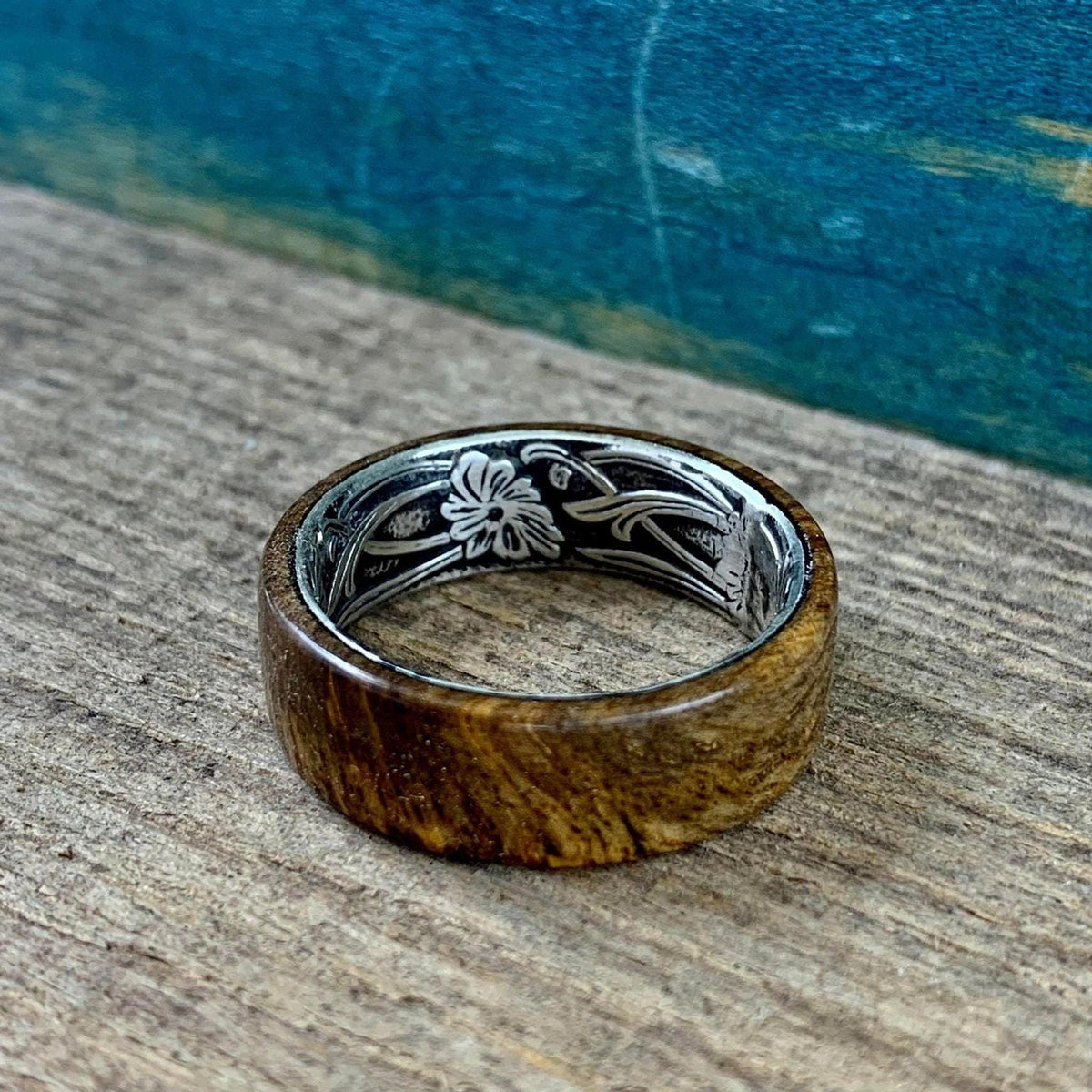 A wooden ring with a silver-colored interior band, engraved with a floral pattern, is shown from a slightly elevated angle on a wooden surface.