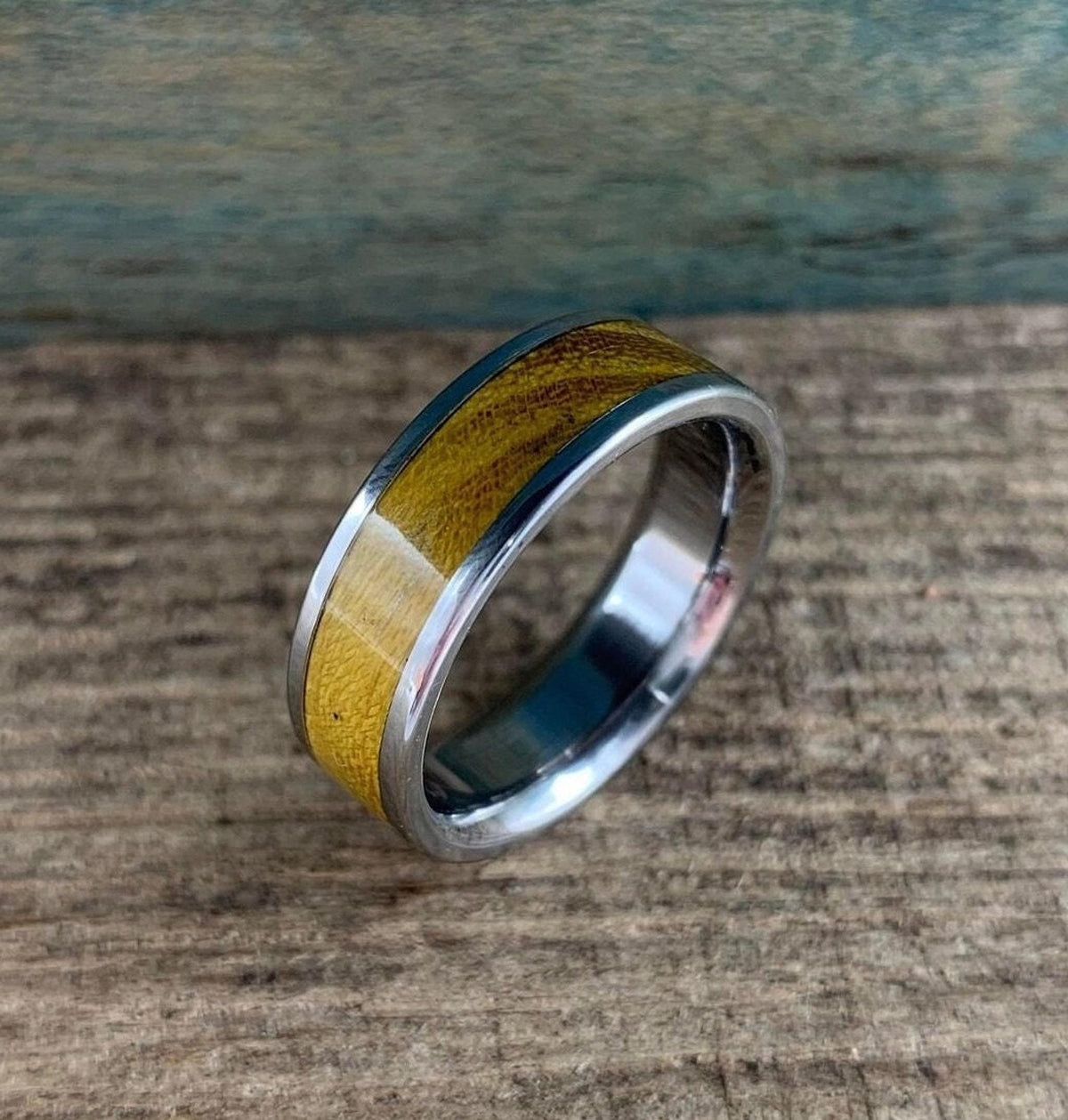 A silver-colored ring with a golden-brown wood inlay is centered on a rustic wooden surface, with a blue-green background.