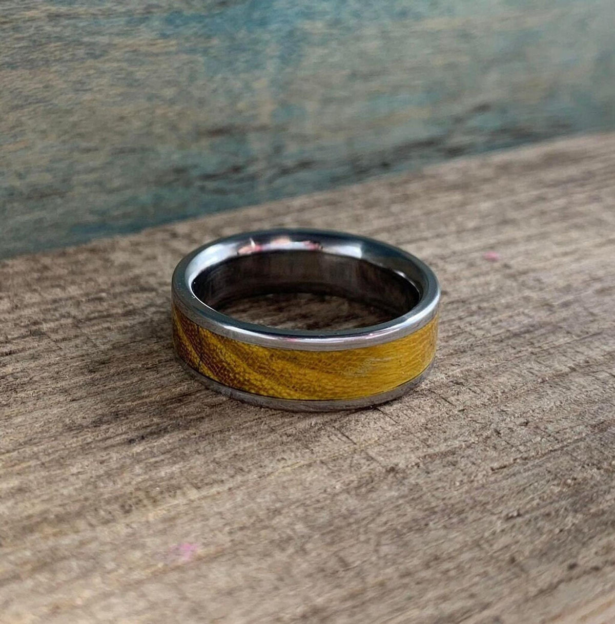 A silver-colored ring with a golden-brown wood inlay is shown from a slightly elevated perspective on a rustic wooden surface.