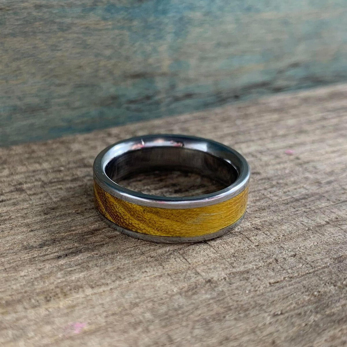A silver-colored ring with a golden-brown wood inlay is centered on a rustic wooden surface, with a blue-green background.