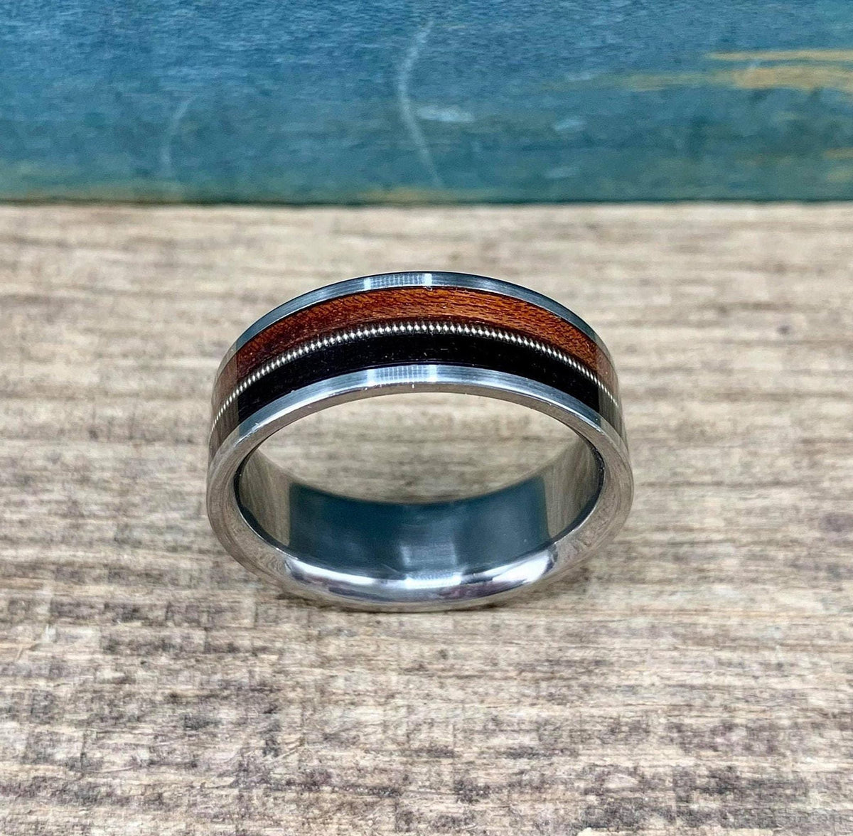 A silver ring with a central band of dark wood, a thin silver guitar string, and a band of reddish-brown wood, is shown from a slightly elevated angle on a light wooden surface.