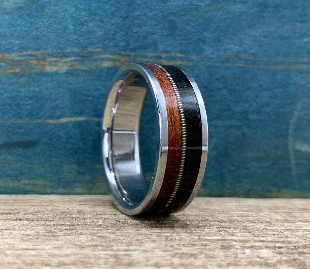 A silver ring with a central band of dark wood, a thin silver guitar string, and a band of reddish-brown wood, is viewed from a side angle on a light wooden surface.
