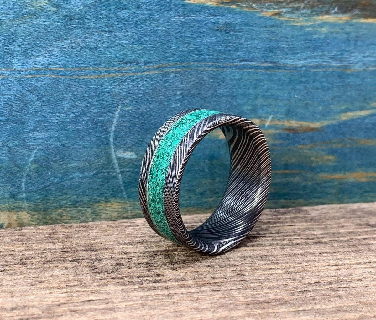 A Damascus steel ring with a malachite inlay, angled on a wooden surface with a blue background.