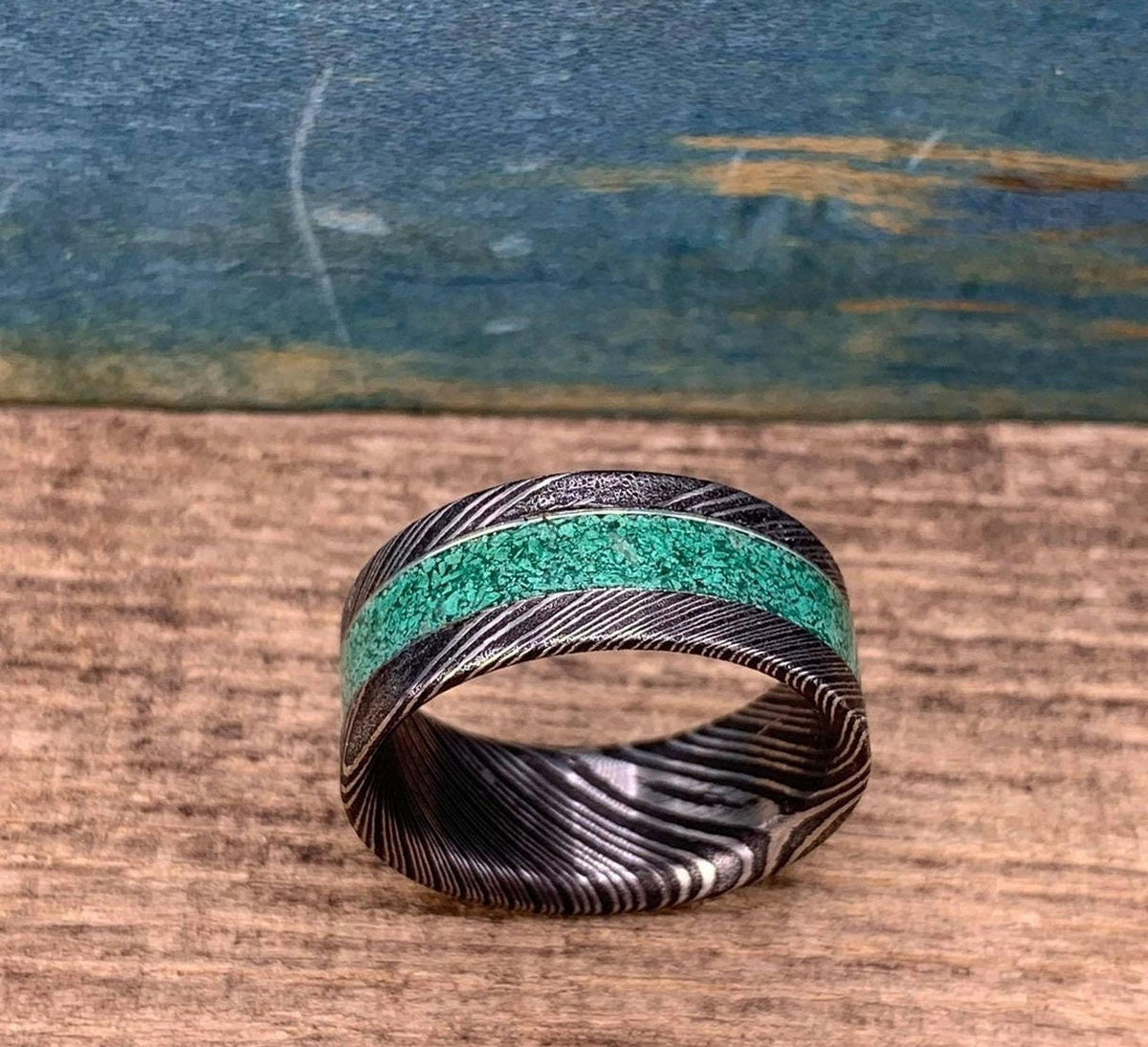 A Damascus steel ring with a malachite inlay, viewed from a slightly different angle on a wooden surface.