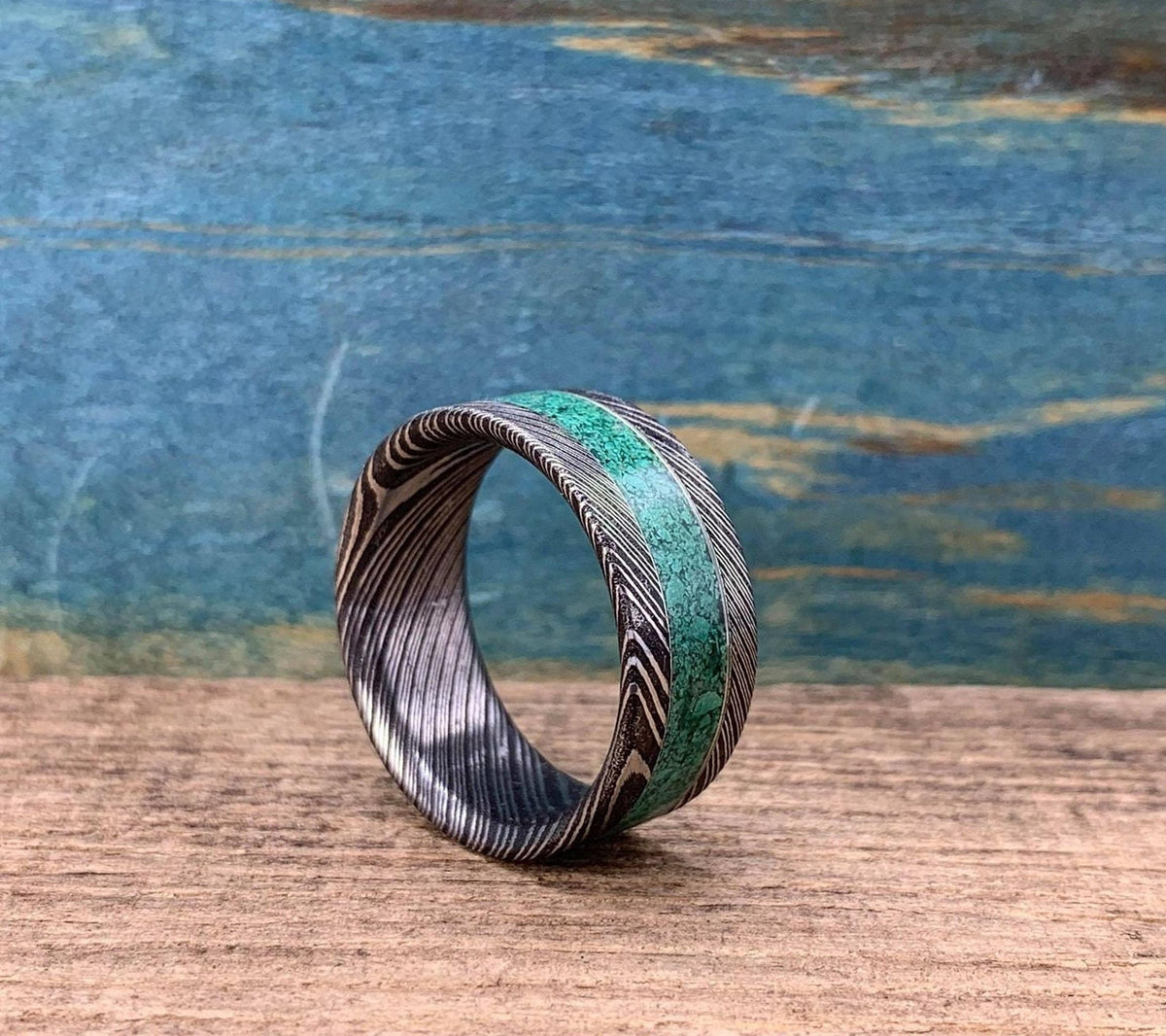 A Damascus steel ring with a malachite inlay, shown from the side on a wooden surface with a blue background.