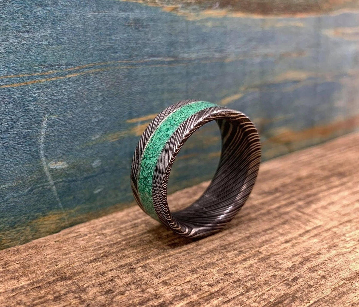 A Damascus steel ring with a malachite inlay, positioned on a wooden surface against a blue wall.