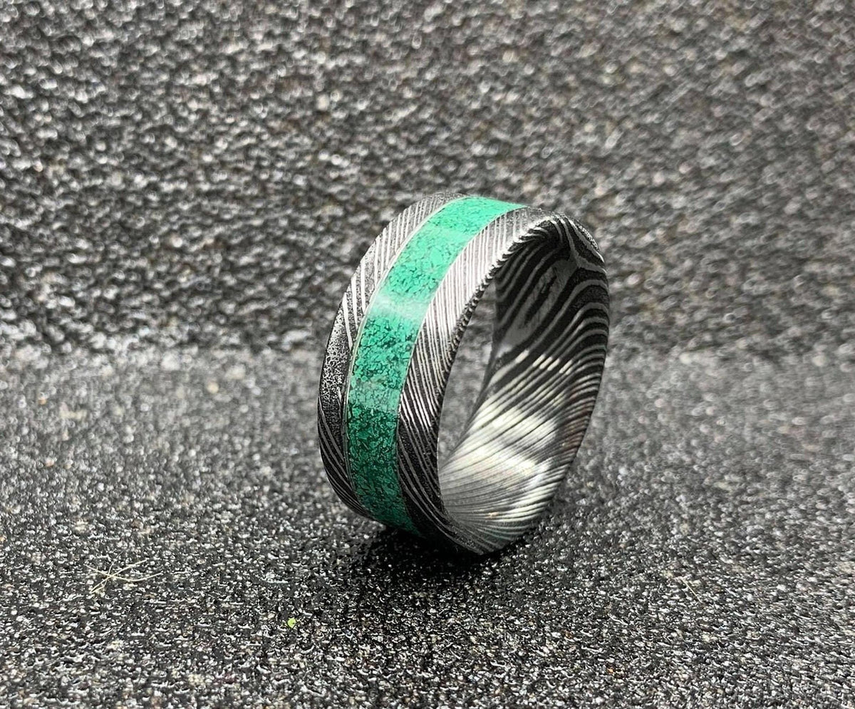 A Damascus steel ring with a malachite inlay, viewed from a slightly elevated angle on a dark, textured surface.