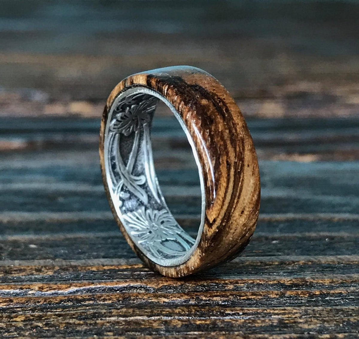 A zebrawood ring with a silver interior featuring an engraved floral pattern is angled on a dark wooden surface.