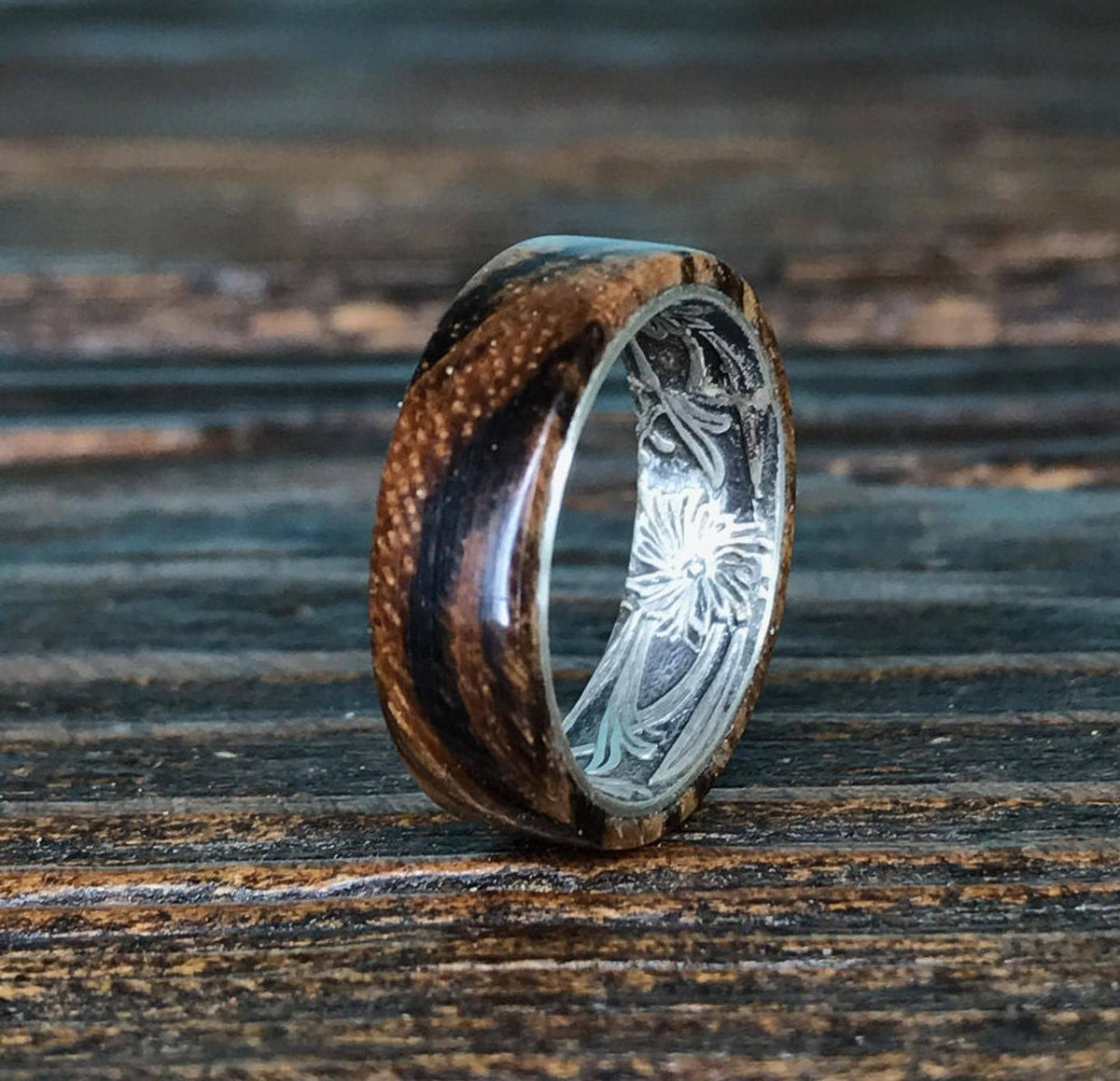 A zebrawood ring with a silver interior and engraved floral pattern is shown from a slightly different angle on a dark wooden surface.