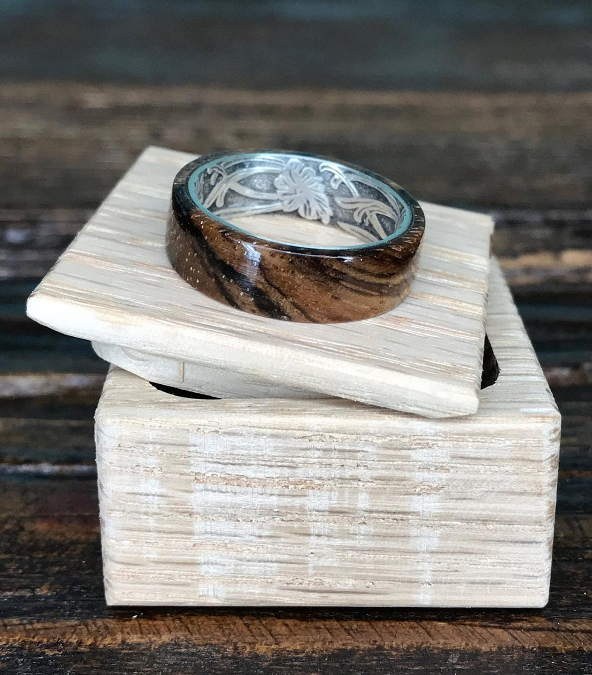 A zebrawood ring with a silver interior and engraved floral pattern rests on a light-colored wooden box with the lid slightly ajar.